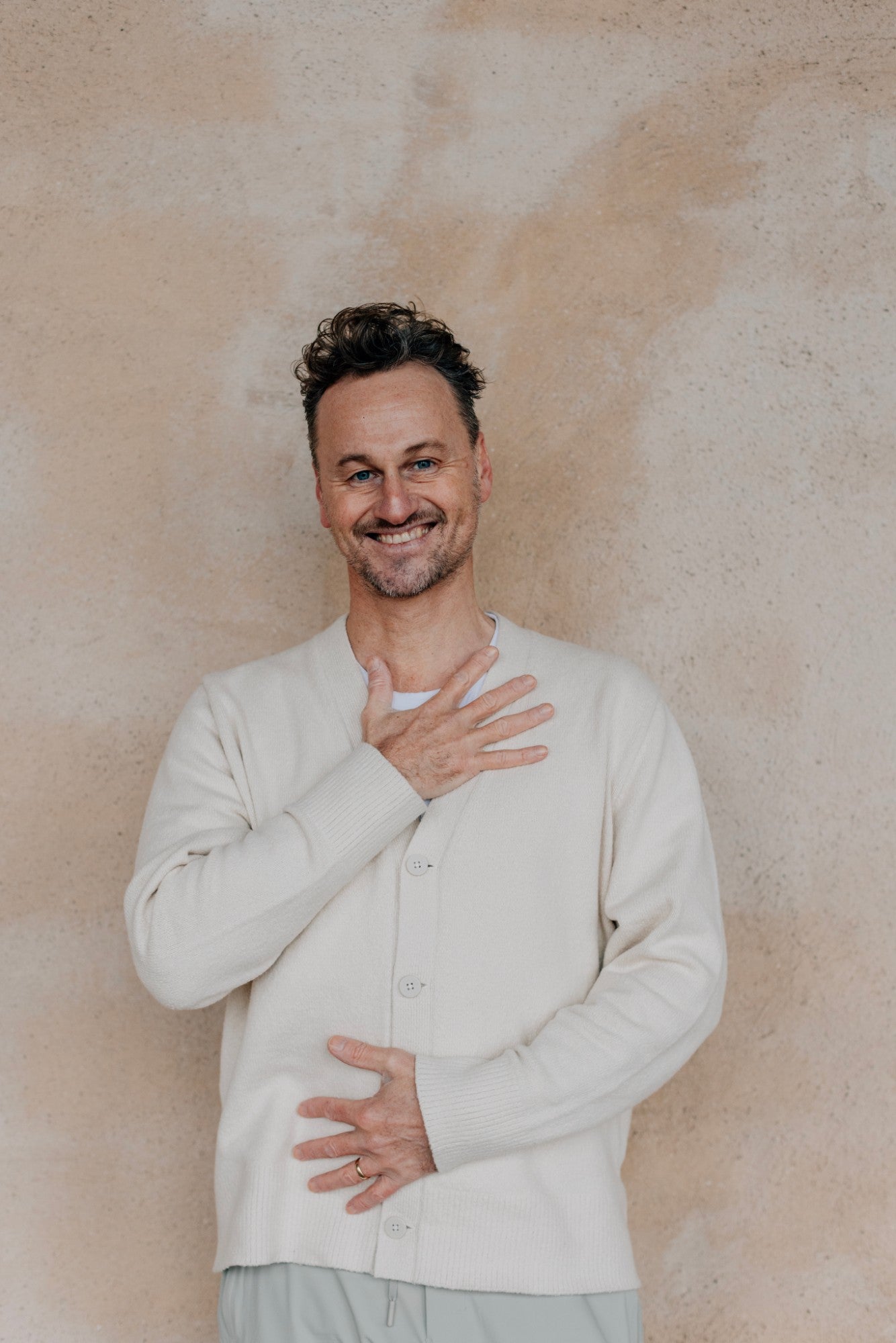 Magnus Fridh ler mot kameran med en hand mot hjärtat, framför en ljus putsad vägg